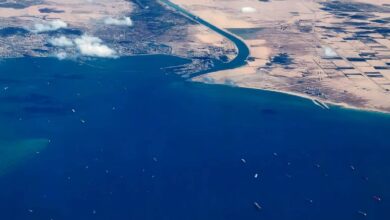 Aerial view of cargo ships queued in the Gulf of Suez near the entrance to the Suez Canal, with desert landscapes and scattered clouds above.