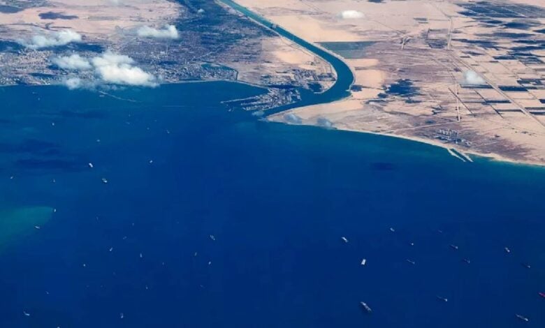 Aerial view of cargo ships queued in the Gulf of Suez near the entrance to the Suez Canal, with desert landscapes and scattered clouds above.