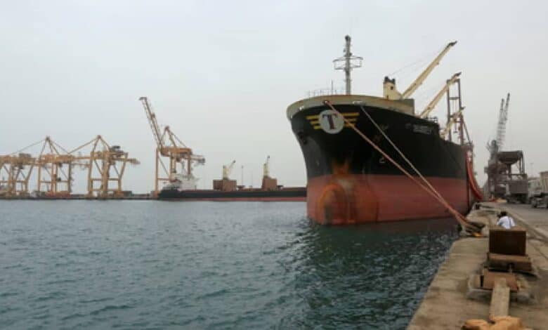 A large cargo ship docked at Hodeidah port, with cranes and other ships in the background, and a worker standing on the dock