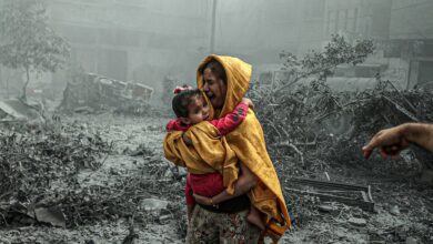 The image of a distressed woman in a yellow shawl carries a frightened child through rubble-filled streets after an Israeli airstrike in Gaza City.