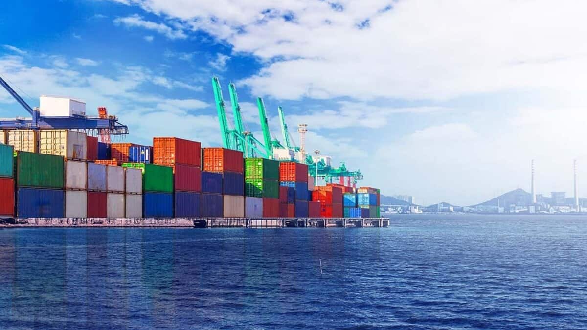 Stacked shipping containers at a busy seaport with cranes by the water.