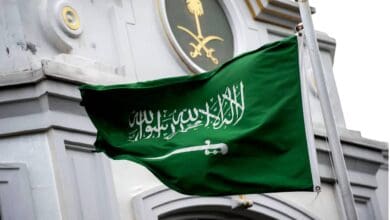 The national flag of Saudi Arabia waves in the wind in front of a government building adorned with the Saudi emblem featuring crossed swords and a palm tree.