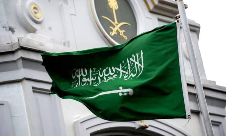 The national flag of Saudi Arabia waves in the wind in front of a government building adorned with the Saudi emblem featuring crossed swords and a palm tree.