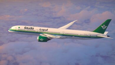 A Saudia airline aeroplane flying above the clouds during sunset, featuring a white fuselage with green and blue stripes, a green tail with a palm tree and two swords logo, and Arabic and English text on the side.