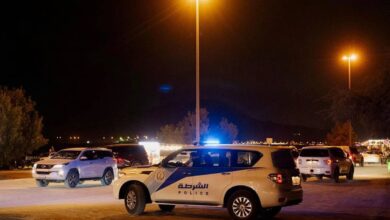 The image of a Sharjah Police patrol vehicle with flashing lights parked at night, surrounded by other cars under streetlights.