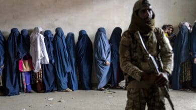 A Taliban fighter in camouflage holding a rifle stands guard in a room, while a line of women in blue burqas waits to receive food rations, with scattered debris on the floor, in Kabul, Afghanistan, May 23, 2023.