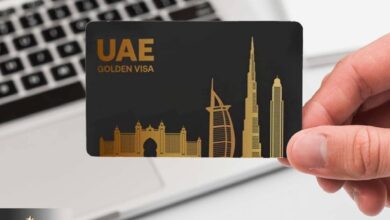 A hand holding a black UAE Golden Visa card with gold-colored silhouettes of iconic buildings, including the Burj Al Arab and Burj Khalifa, set against a laptop keyboard in the background.
