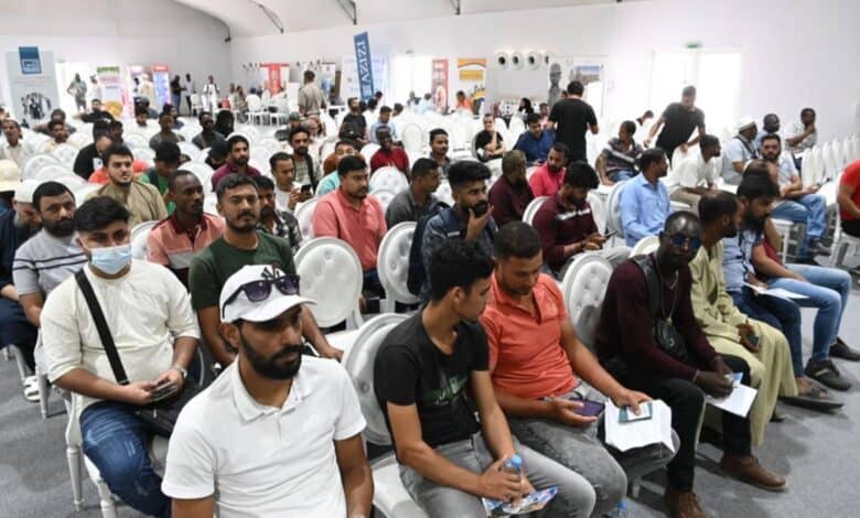 Group of people seated in a conference hall, holding documents, with banners in the background, likely related to UAE visa violation processing.