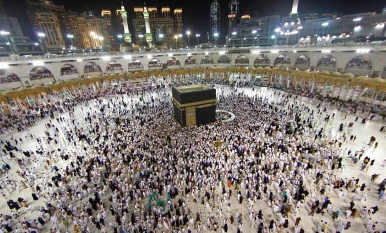 A large group of Muslim worshippers, dressed in white Ihram garments, perform Tawaf by circling the Kaaba at the center of Grand Mosque in Makkah, Saudi Arabia, during the Umrah pilgrimage at night. The illuminated minarets and architecture of the holy mosque create a vibrant skyline, reflecting the spiritual reverence and unity of the gathered devotees.