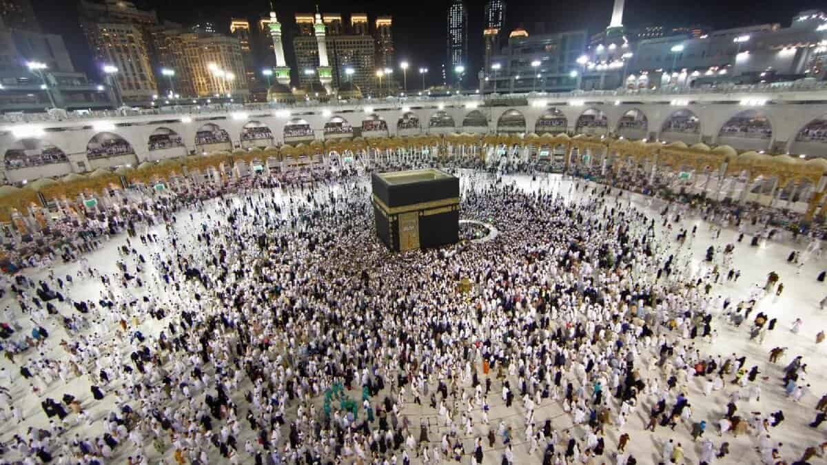 A large group of Muslim worshippers, dressed in white Ihram garments, perform Tawaf by circling the Kaaba at the center of Grand Mosque in Makkah, Saudi Arabia, during the Umrah pilgrimage at night. The illuminated minarets and architecture of the holy mosque create a vibrant skyline, reflecting the spiritual reverence and unity of the gathered devotees.