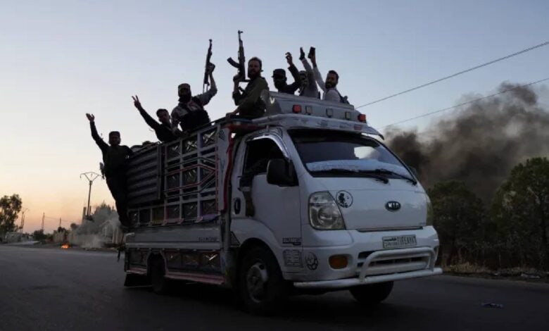 A group of armed Bedouin fighters raise their weapons while riding on a white pickup truck along a street in Sweida province, with black smoke rising in the background during ongoing sectarian clashes.