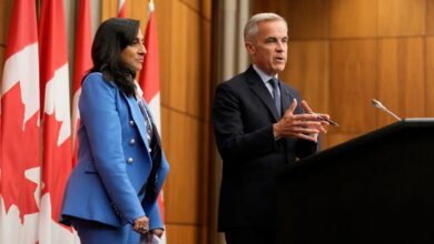 Canadian Prime Minister Mark Carney speaks at a podium during a press conference, accompanied by a senior official, with Canadian flags in the background.
