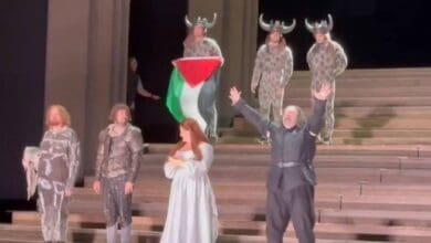 scene from a performance at the Royal Opera House where a cast member unfurls a Palestinian flag, surrounded by other performers in costumes, including some wearing horned helmets, on a tiered stage.