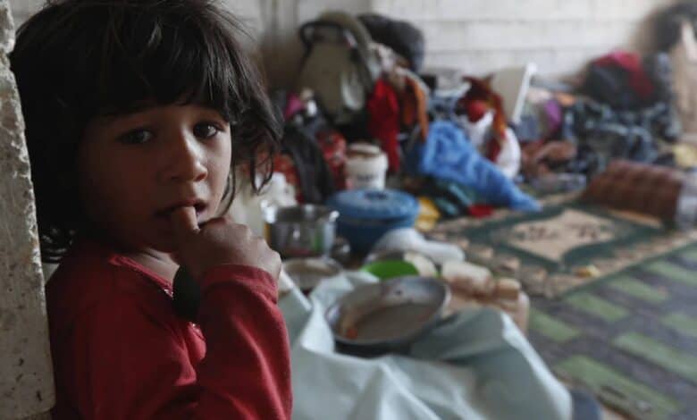 A displaced Bedouin girl in a red shirt stands inside a bare shelter, looking at the camera, with scattered household items and bedding behind her.