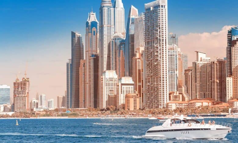 A luxury yacht cruises across the blue waters in front of Dubai Marina’s modern skyscrapers, under a clear sky.