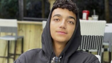 Edan Alexander, wearing a black hoodie and sunglasses around his neck, sits outdoors at a cafe.
