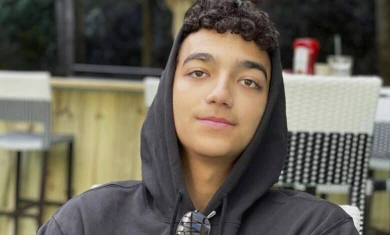 Edan Alexander, wearing a black hoodie and sunglasses around his neck, sits outdoors at a cafe.