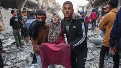 Two men carry an injured elderly woman through the rubble of destroyed buildings in Gaza, as others look on amidst the devastation.