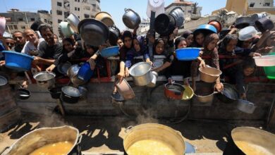 Palestinians crowd around with empty pots and containers, reaching over a barrier to receive food from steaming cauldrons during a mass distribution in Gaza. The scene reflects the severe humanitarian crisis and widespread hunger in the region.