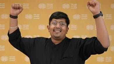 A joyful man with raised arms, wearing a black shirt and glasses, standing against a yellow patterned background.