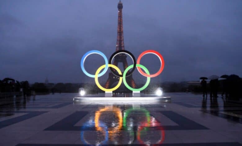 Olympic rings displayed on Trocadero Esplanade with Eiffel Tower in the background, wet pavement reflecting the rings under a rainy sky.