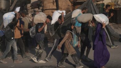 A group of Palestinians, covered in dust, walk through rubble-strewn streets carrying heavy sacks of flour on their shoulders. Some wear backpacks, and a woman in a purple robe is seen in the front. The aid is part of a humanitarian convoy that reached Gaza City.