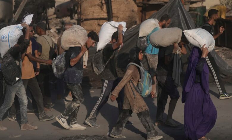 A group of Palestinians, covered in dust, walk through rubble-strewn streets carrying heavy sacks of flour on their shoulders. Some wear backpacks, and a woman in a purple robe is seen in the front. The aid is part of a humanitarian convoy that reached Gaza City.