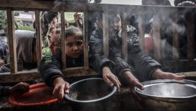 Palestinian children and adults, dressed in jackets, hold out metal pots and plates through a wooden fence, reaching for food amid rising steam in a crowded, desperate scene in Gaza,.