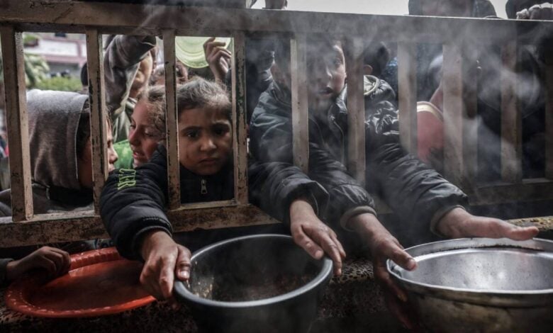 Palestinian children and adults, dressed in jackets, hold out metal pots and plates through a wooden fence, reaching for food amid rising steam in a crowded, desperate scene in Gaza,.