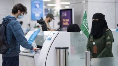 A Saudi female immigration officer in uniform processes a male passenger at an airport immigration counter.