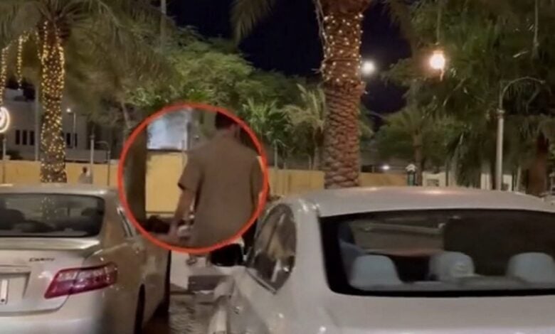 A man circled in red is seen walking between parked cars at night in a palm-lined street, reportedly wearing a night thobe. The scene is dimly lit with streetlights and decorative tree lights.