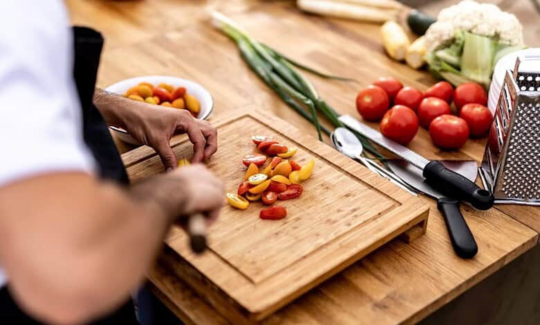 Chef chopping fresh cherry tomatoes on a wooden board with vegetables and kitchen tools around.