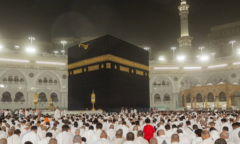 Pilgrims in white Ihram gather at the Grand Mosque in Makkah, surrounding the Kaaba in prayer under bright lights during a sacred moment of worship.