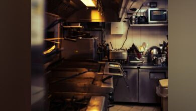 A busy kitchen with stainless steel appliances, a microwave, pots, pans, and a sink, illuminated by warm lighting.
