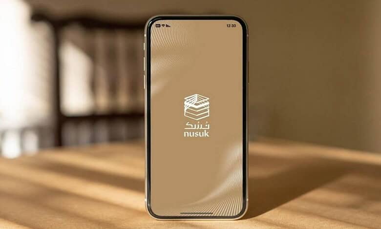 The image of a smartphone on a wooden table showing the Nusuk app splash screen with Arabic logo on a beige background.