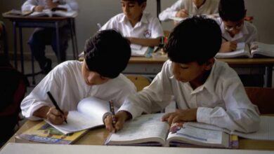 Two students in white uniforms writing in notebooks at desks in a UAE school classroom, with other students working in the background.