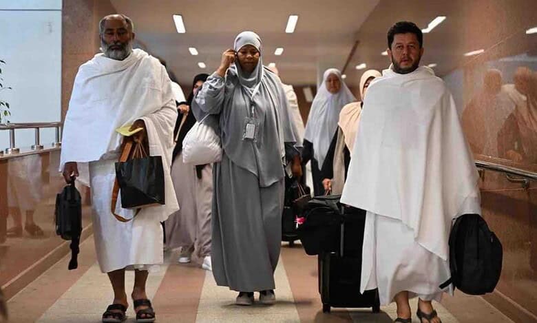 A group of Umrah pilgrims, dressed in Ihram and modest attire, walking through an airport corridor in Saudi Arabia. Some carry luggage and bags, with a modern interior featuring glass railings and overhead lighting.