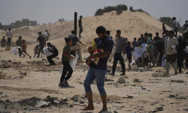 A crowded scene of people, including a man holding a child, walking across a debris-strewn sandy area, seeking aid in a conflict zone.