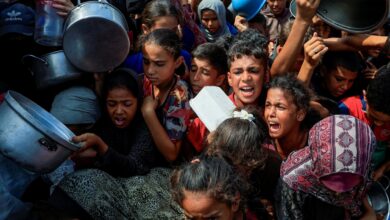 A crowded scene of people, mostly children, holding pots and containers, reaching out in a desperate manner.