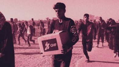 A young Palestinian man carries a Gaza Humanitarian Foundation (GHF) aid box while others walk and run behind him in a crowded, dusty area.