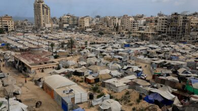 A sprawling refugee camp in Gaza with numerous tents and makeshift shelters, surrounded by damaged buildings and a tall, partially destroyed high-rise, under a cloudy sky.
