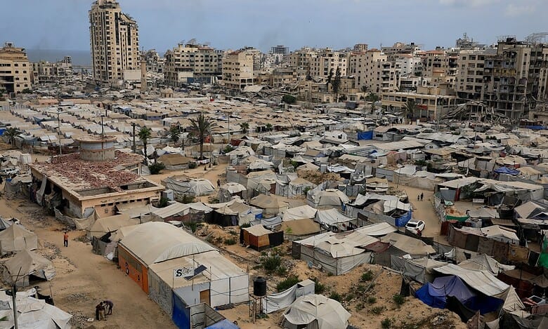 A sprawling refugee camp in Gaza with numerous tents and makeshift shelters, surrounded by damaged buildings and a tall, partially destroyed high-rise, under a cloudy sky.