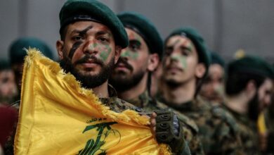 A Hezbollah militant holds the group's yellow flag during a parade, with fellow fighters in camouflage and green berets standing behind him.