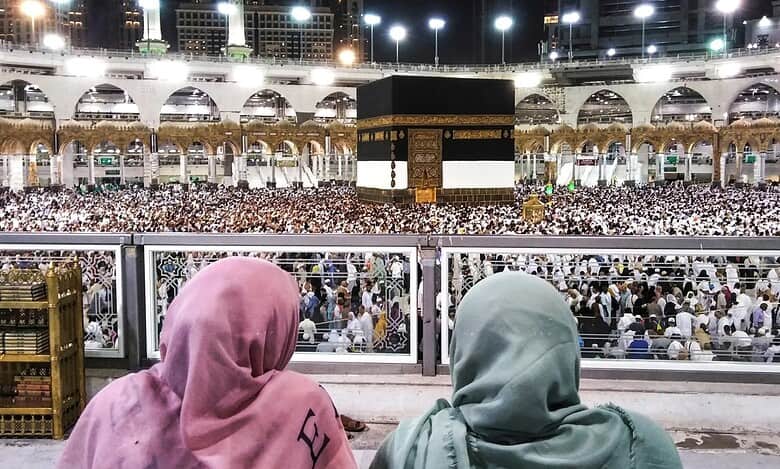 Two people in colorful headscarves observe a large crowd gathered around the Kaaba at night in a grand mosque.