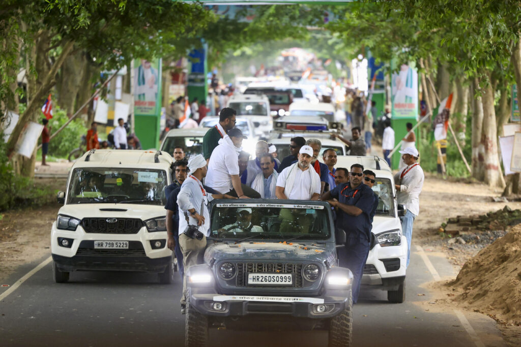 Busy political rally procession in India with security and supporters on a tree-lined road.