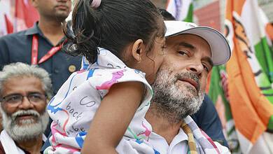 Child whispering to man at political rally, India election campaign, vibrant flags in background.