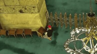 Overhead view of a person in a red cap and dark clothing near a yellow structure, with chairs and a chandelier in the background, inside a mosque.