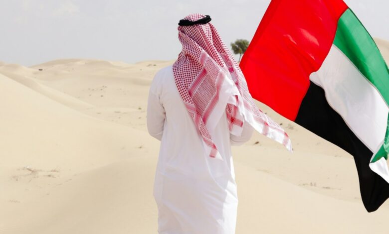 Man in traditional Emirati attire holding UAE flag in desert.