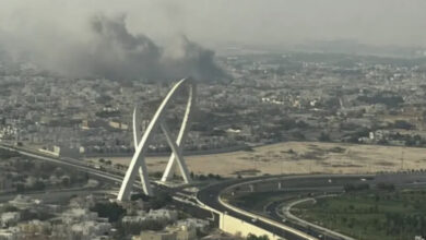 Smoke rises over buildings in Doha near a landmark arch.