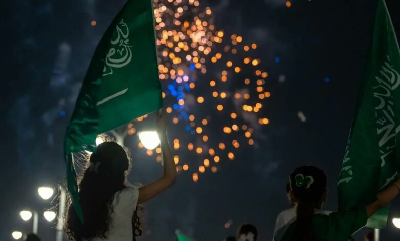 Silhouettes of children waving Saudi flags against a night sky filled with colorful fireworks, celebrating the 95th National Day, with streetlights illuminating the scene below.